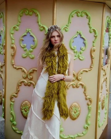 A woman with long blonde hair poses in a white dress with a green scarf against a decorative wall with pink, green, and gold details.