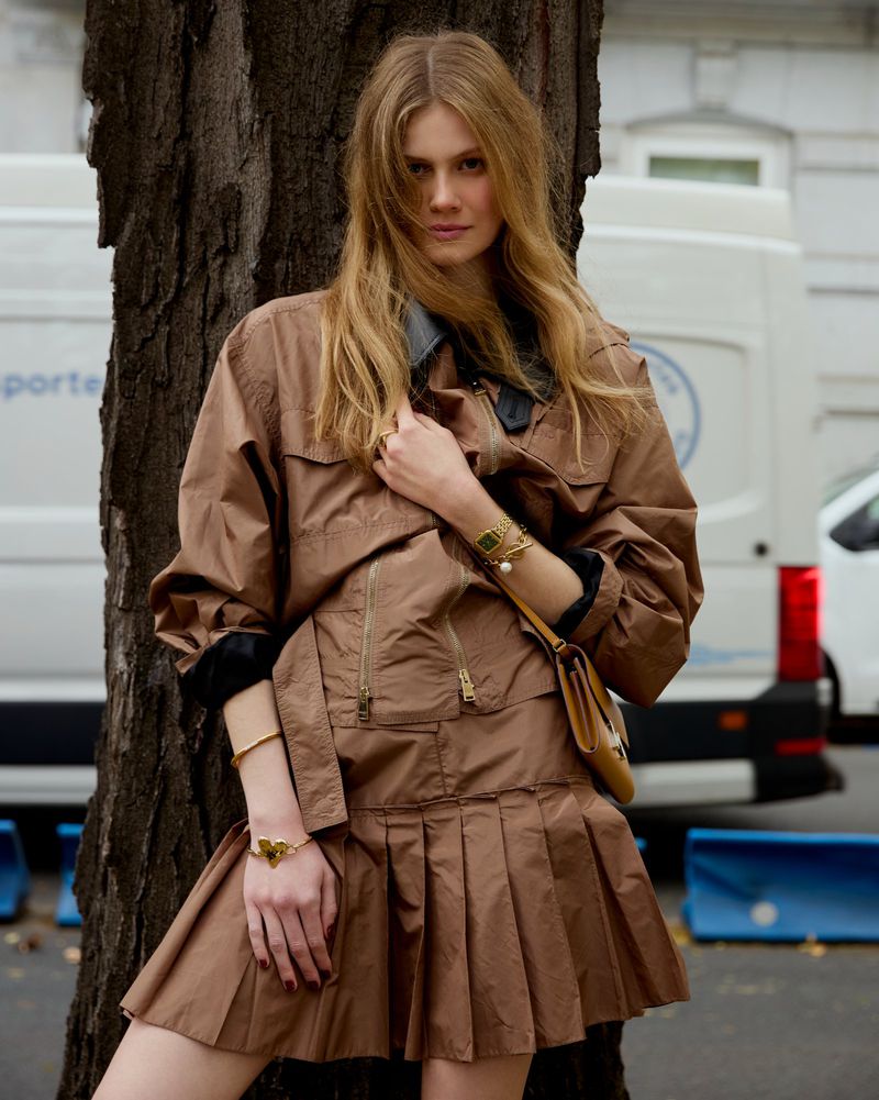 Young woman in brown jacket and skirt standing near a tree next to a white van with blue accents in an urban environment.