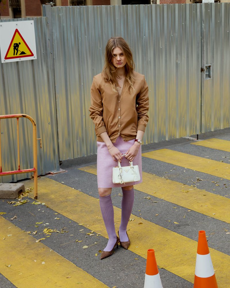 Woman in brown cardigan and pink skirt poses near metal construction barrier with handbag and knee-high socks on a striped road.