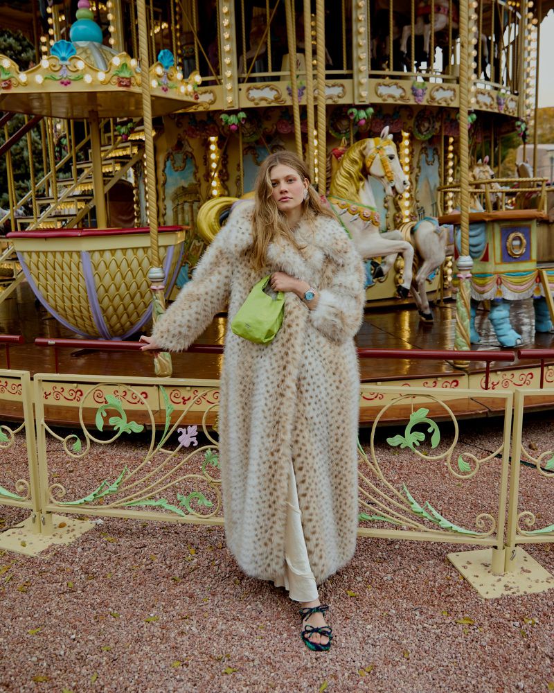 A woman in a faux fur coat stands in front of a carousel. She is holding a green bag and wearing sandals.