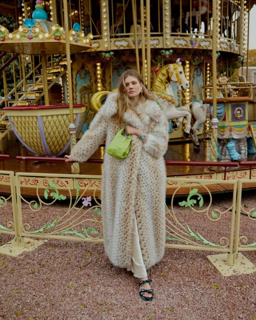 A woman in a faux fur coat stands in front of a carousel. She is holding a green bag and wearing sandals. 