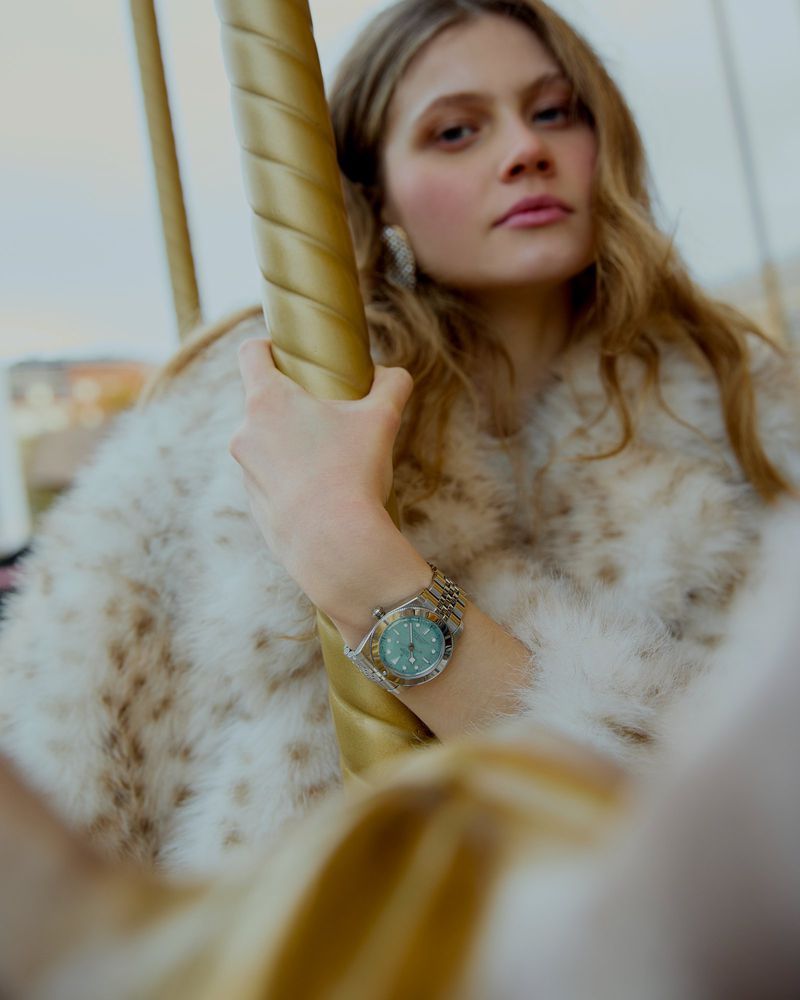 Blonde woman wearing a fur coat and a watch posing on a carousel with a fairground ambiance, soft lighting, and a focus on fashion.