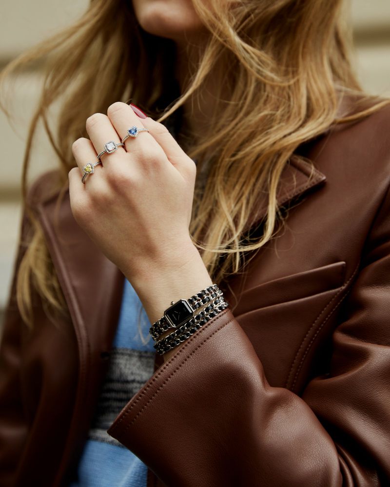 Close-up of woman's hands with three rings, brown leather jacket, and wristwatch with dark band, set against an outdoor backdrop.