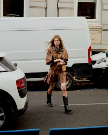 An adult woman walks on a city street, wearing a brown jacket and skirt, black boots, and carrying a small purse, with white cars nearby.