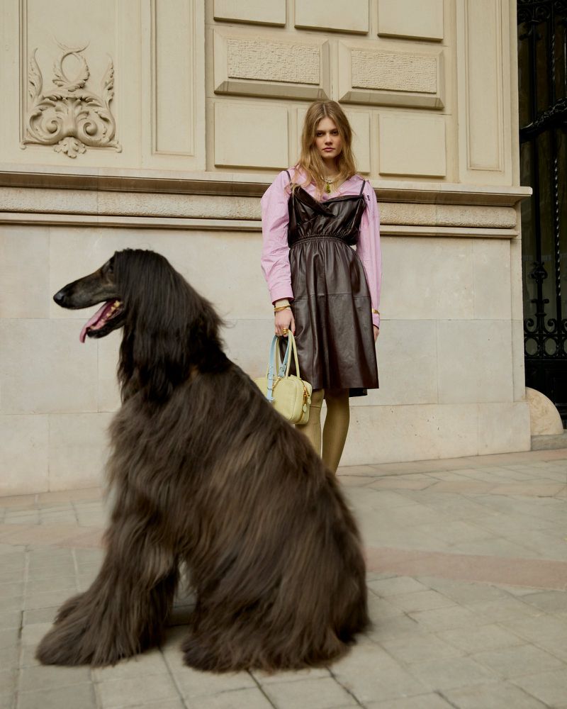 Woman in a brown dress posing outside with an Afghan Hound dog; the building has decorative elements in the background.