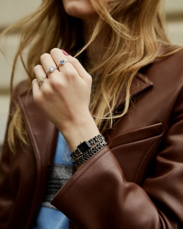 Woman in a brown leather jacket showcasing a black double-wrap chain wristwatch and gemstone rings on her fingers.