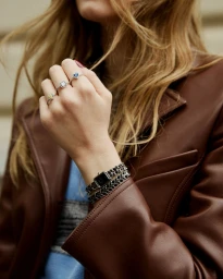 Woman in a brown leather jacket showcasing a black double-wrap chain wristwatch and gemstone rings on her fingers.