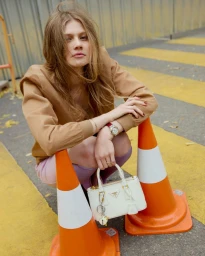 A woman with wavy hair crouches between two orange traffic cones on a street holding a small white designer handbag.