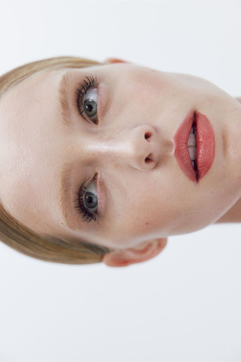 Close-up portrait of a fair-skinned young woman with light hair and light eyes, wearing light pink lipstick, against a white background.