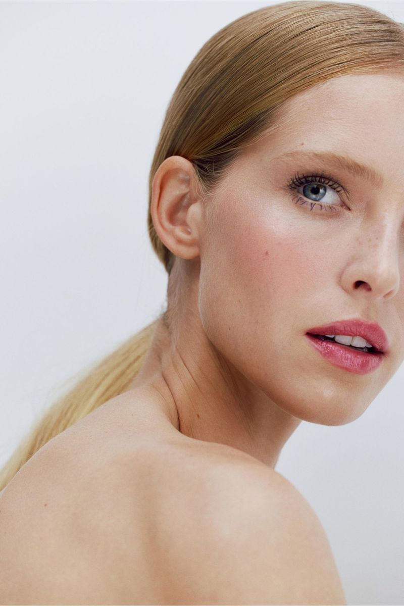 Close-up portrait of a young adult woman with blonde hair pulled back in a ponytail, fair skin, and pink lipstick against a white background.