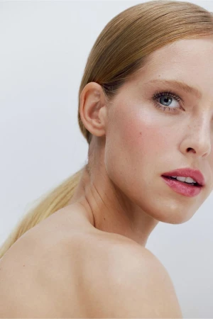Close-up portrait of a young adult woman with blonde hair pulled back in a ponytail, fair skin, and pink lipstick against a white background.