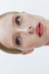 Close-up portrait of model Abby Champion with natural makeup and coral lipstick against a plain white background.