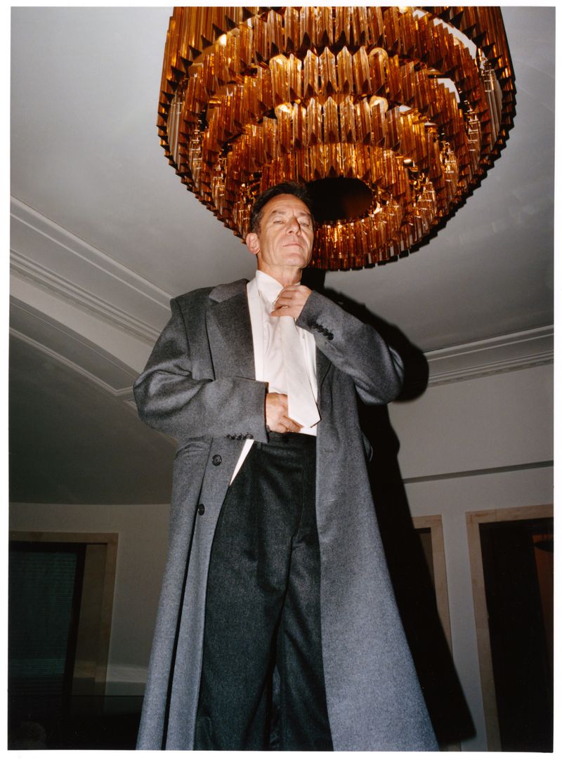 Stylish man in grey coat adjusting collar, standing indoors beneath a large gold chandelier, looking at the camera.