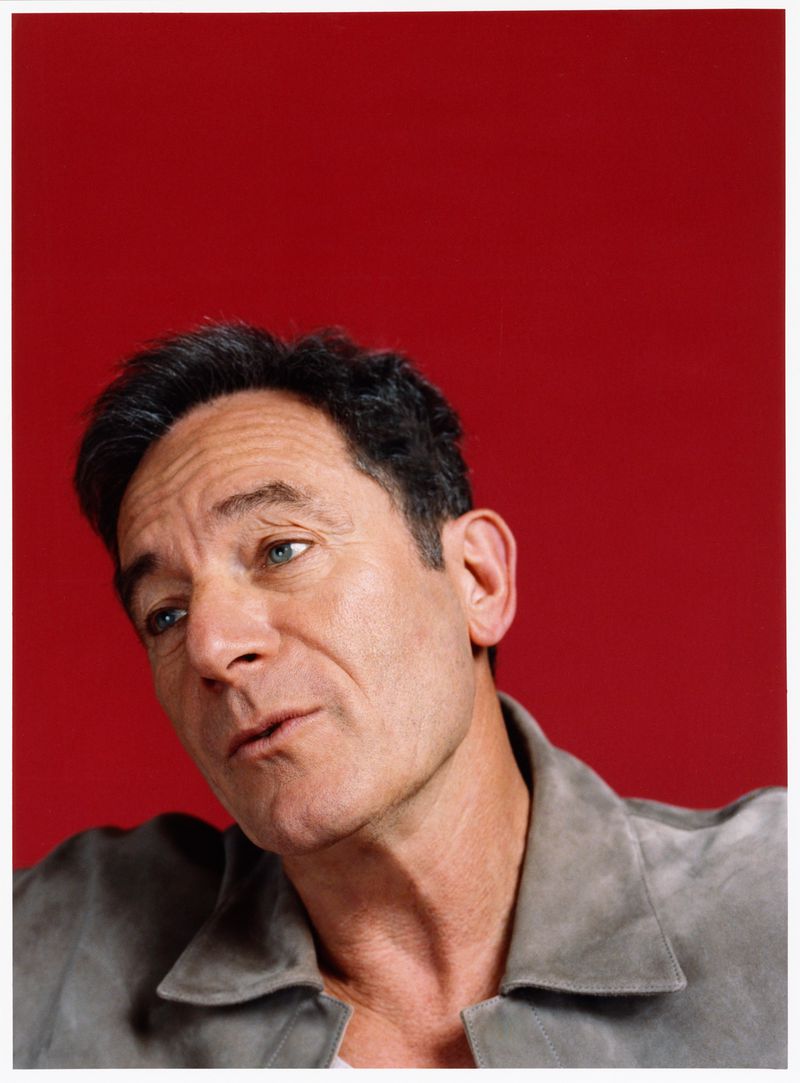 Portrait of a mature adult male with dark hair wearing a gray jacket, set against a bright red background in a photo studio.