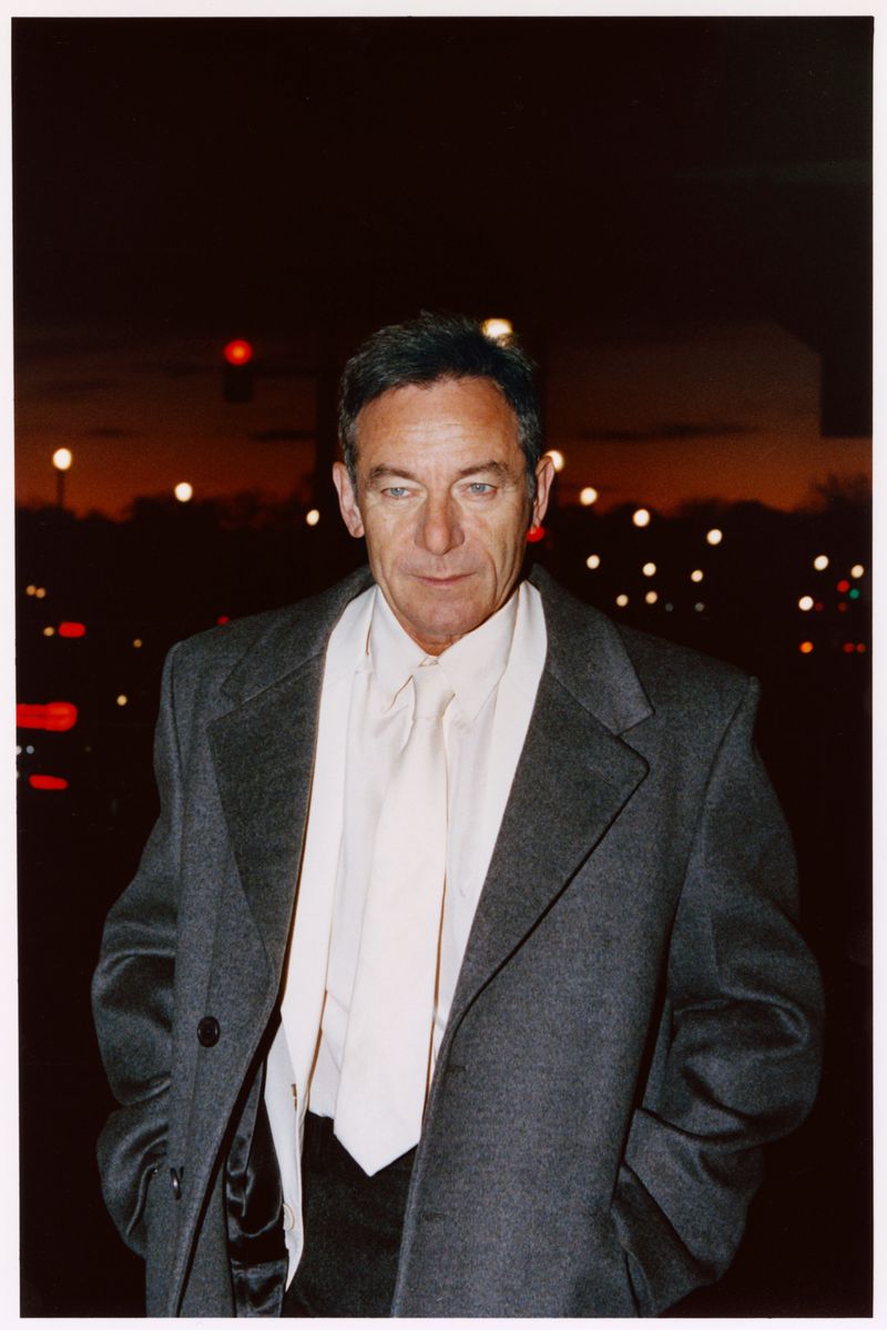 Headshot of an adult man wearing a gray coat and white collared shirt, photographed outdoors at night with blurred lights in the background.