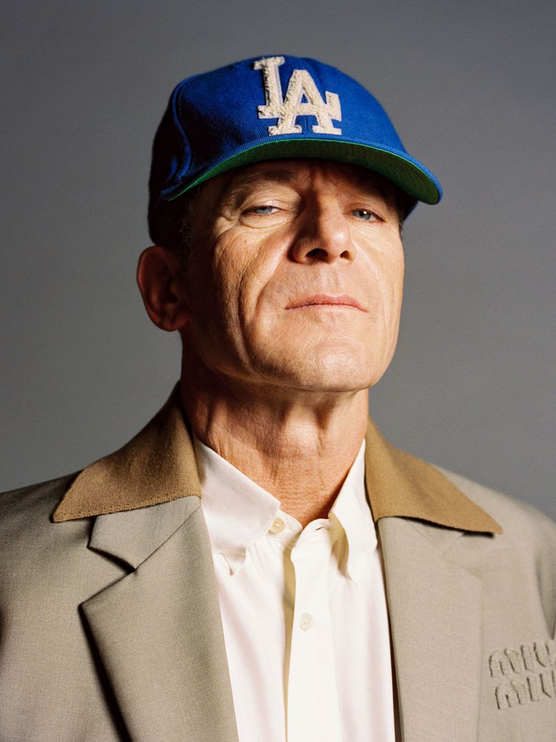 Portrait of an adult man in a beige jacket and blue LA baseball cap against a grey backdrop, shot at eye-level in medium framing.