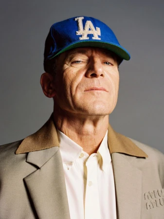 Portrait of an adult man in a beige jacket and blue LA baseball cap against a grey backdrop, shot at eye-level in medium framing.