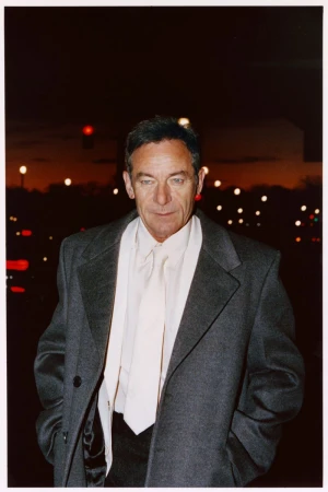 Actor Jason Isaacs wearing a grey coat and white tie outdoors at night with blurred city lights in the background.