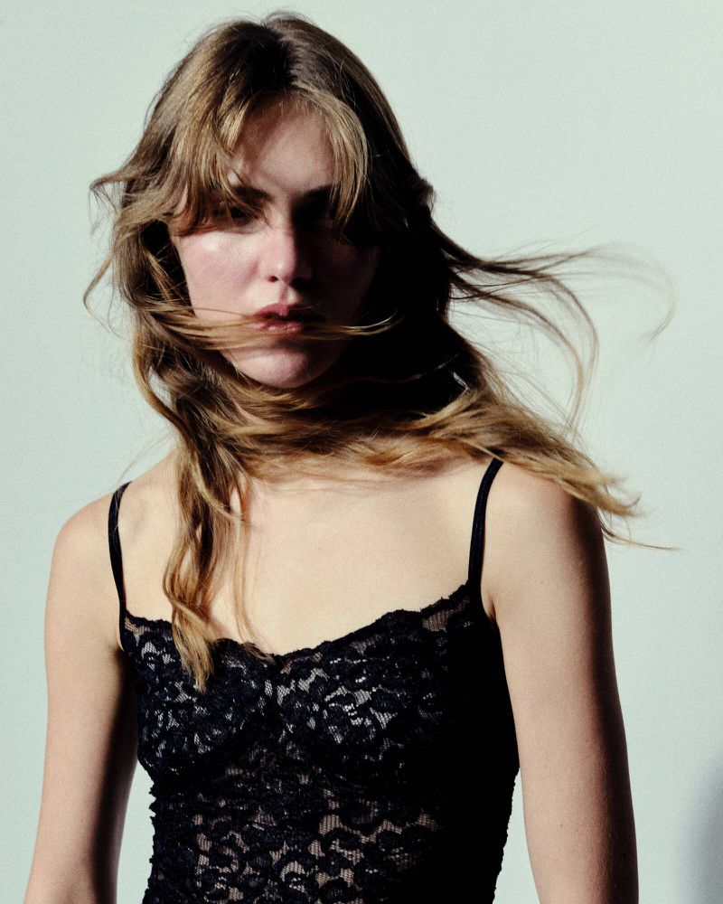 Close-up of a young adult woman with long brown hair wearing a black lace top, posing against a light grey background in a studio.