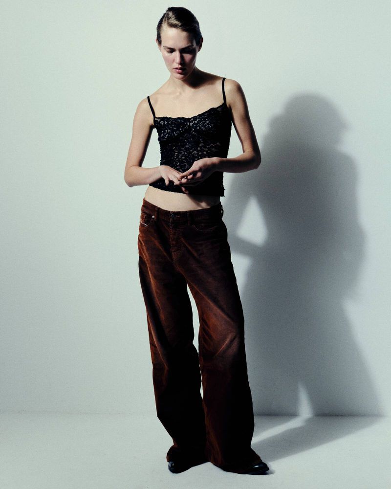 Full-length shot of a woman in a black spaghetti-strap top and brown corduroy pants against a white backdrop with a subtle shadow.
