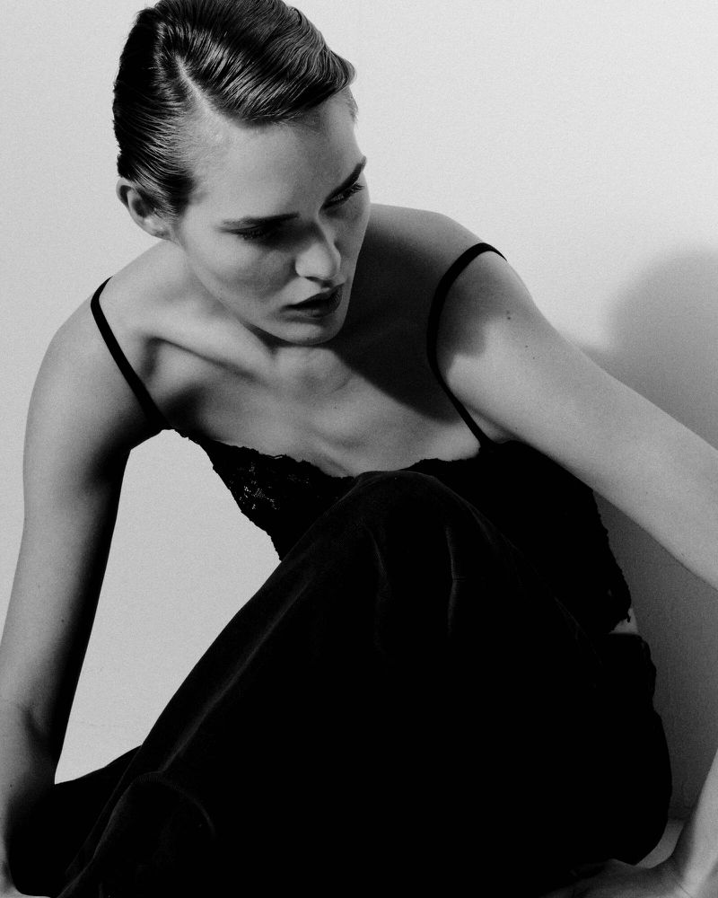 Black and white portrait of a woman with slicked-back hair wearing a camisole, posing in a photo studio.