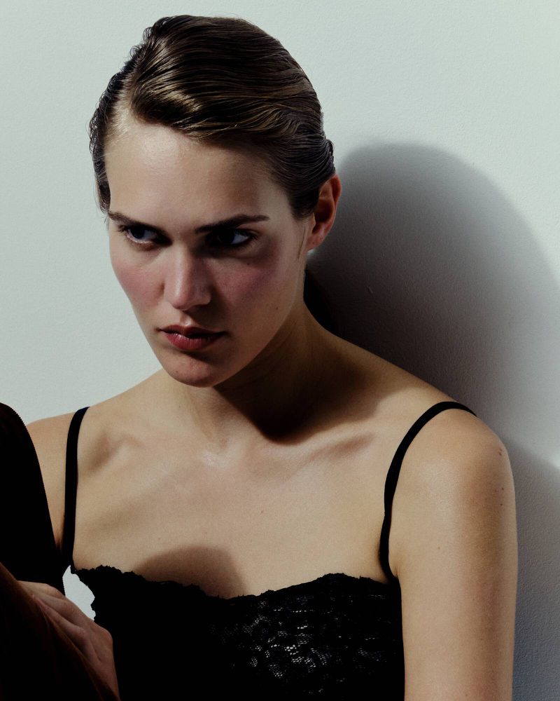 Woman with slicked-back brown hair and black tank top posing against a white wall in a portrait. Fashion photography.