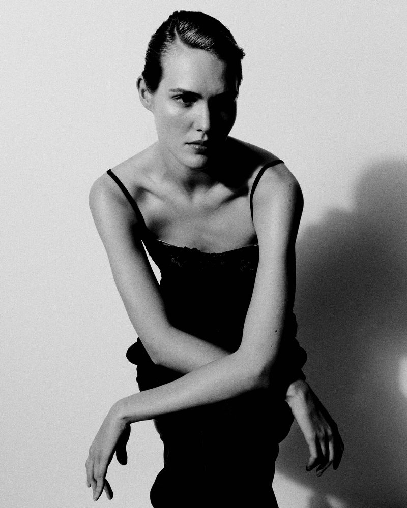 Portrait of a poised adult female fashion model indoors, wearing an elegant dark dress against a white background with soft lighting.