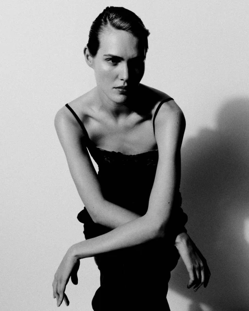 Portrait of a poised adult female fashion model indoors, wearing an elegant dark dress against a white background with soft lighting.