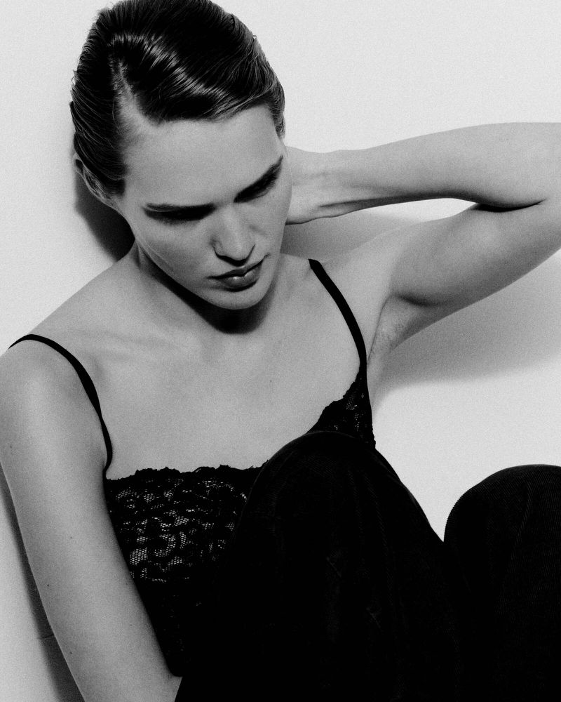 Close-up portrait of a woman in a black lace top with her hand behind her head against a plain white background