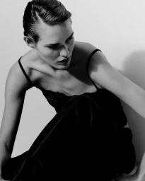 Model with slicked-back hair wearing a black lace camisole and dark trousers crouching against a pale studio wall in black and white.