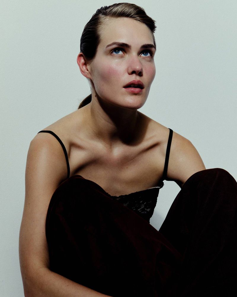 A woman with slicked-back hair wearing a black spaghetti-strap top looks upward against a neutral pale studio backdrop.