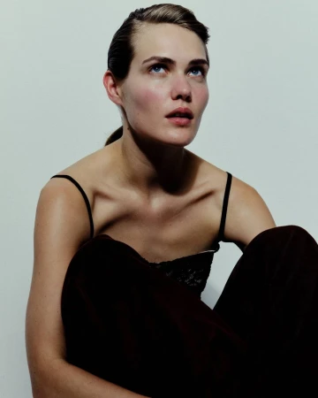 A woman with slicked-back hair wearing a black spaghetti-strap top looks upward against a neutral pale studio backdrop.