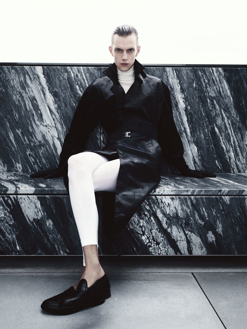 A slender adult male is seated on a bench with dark marble, wearing a long black leather coat, white leggings, and black loafers.
