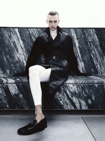 A slender adult male is seated on a bench with dark marble, wearing a long black leather coat, white leggings, and black loafers.
