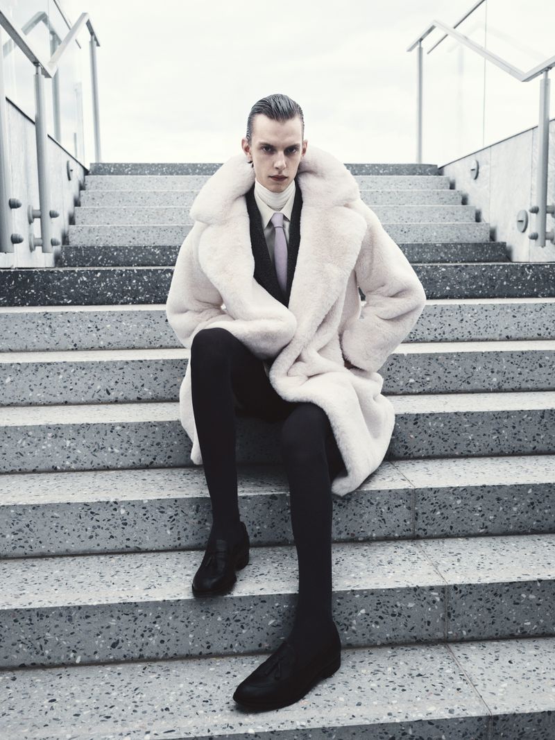Fashionable adult male with slicked-back hair, wearing a luxurious fur coat and black suit, sitting on concrete steps in an urban setting.