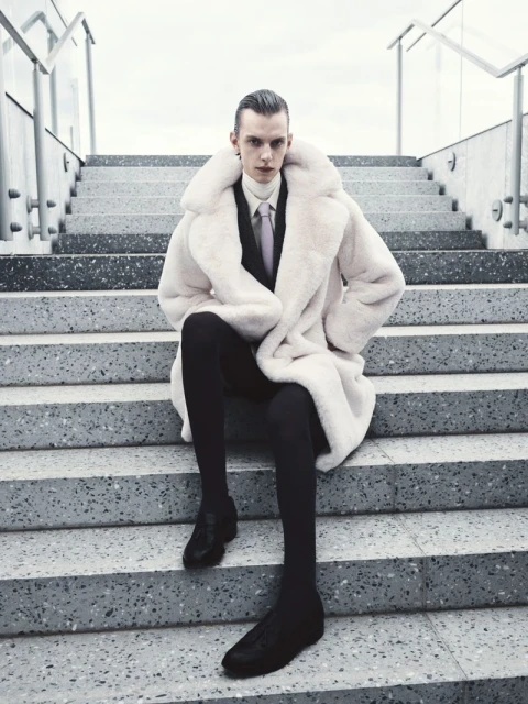 Fashionable adult male with slicked-back hair, wearing a luxurious fur coat and black suit, sitting on concrete steps in an urban setting.