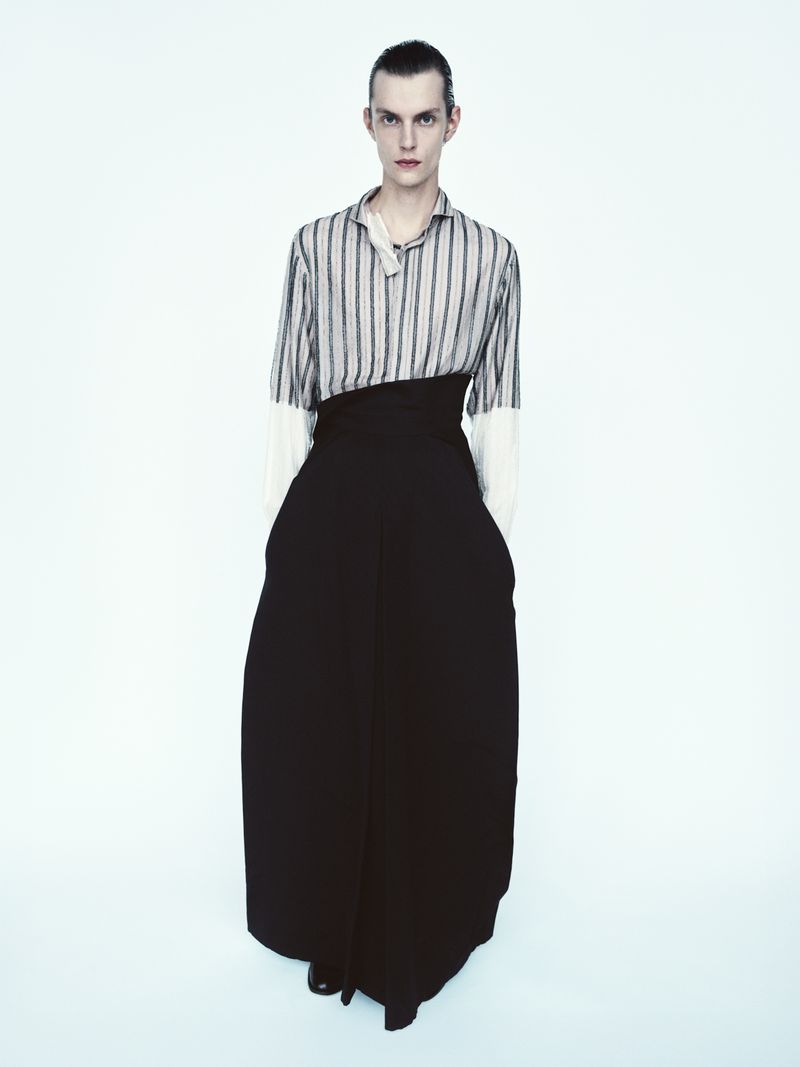 Fashionable person posing in a striped shirt and wide-legged black pants against a white background, presenting an editorial style.