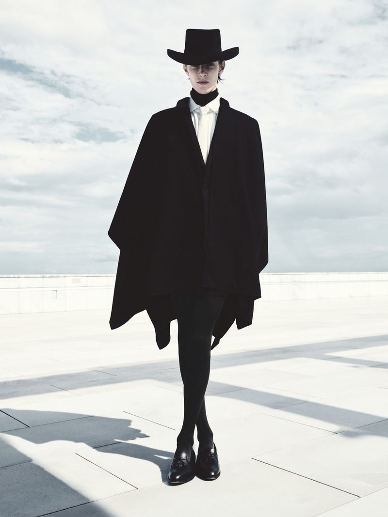 A person stands on a white rooftop wearing a black hat, cape, shirt, tights, and loafers against a cloudy sky background.