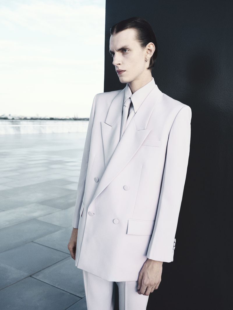 Fashionable adult male in a double-breasted white suit with a necktie, posing against a gradient sky and black wall.