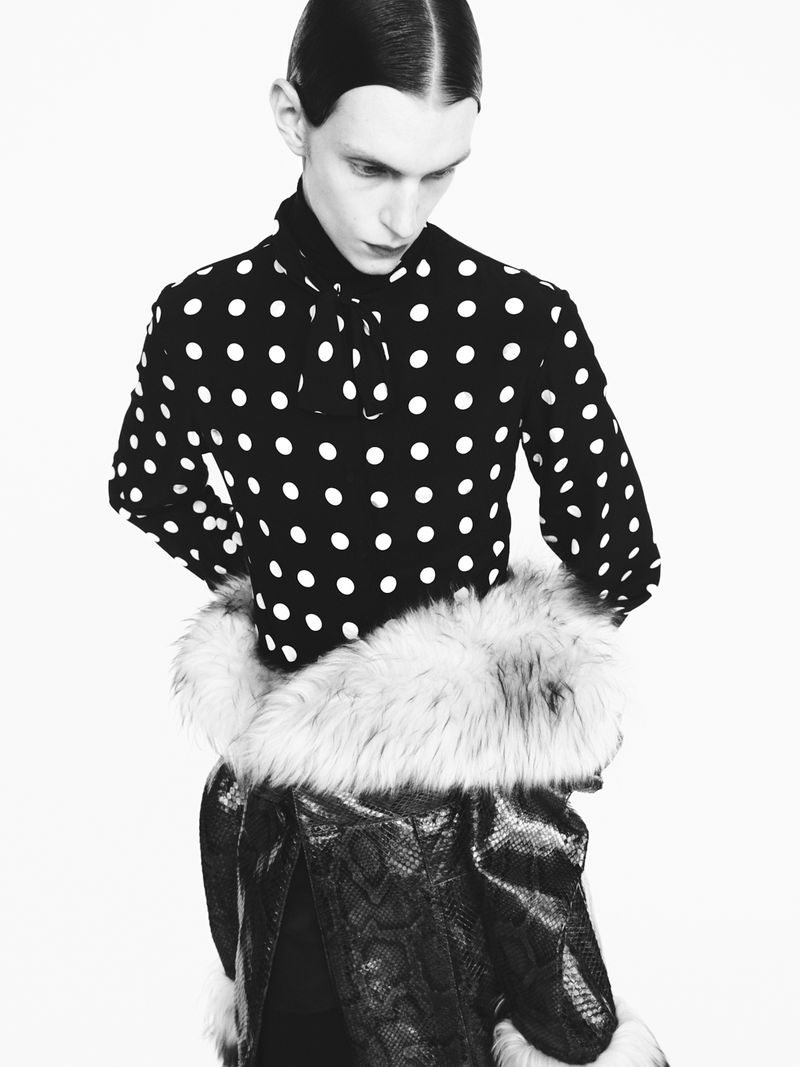 Fashion shot of a slender person wearing a black and white polka dot top, a fur collar, and a black snakeskin pattern skirt against a white background.