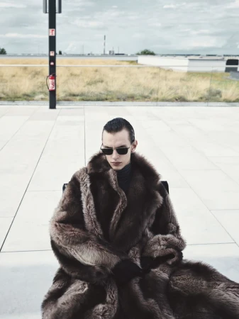 A person in sunglasses and a fur coat sits on a chair outdoors on a paved surface, with buildings and a cloudy sky in the background.