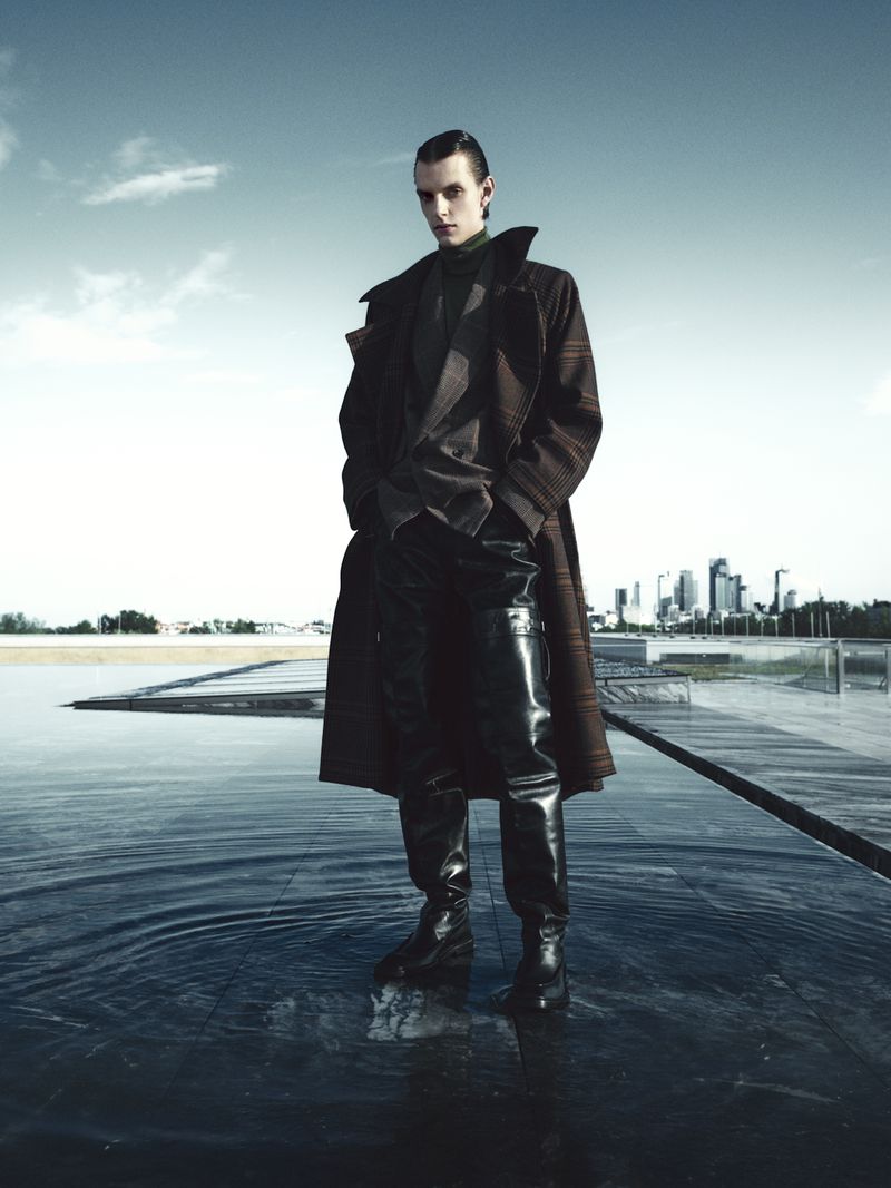 A lone male model strikes a confident pose in an overcoat and black leather pants, set against a modern urban backdrop and still water.