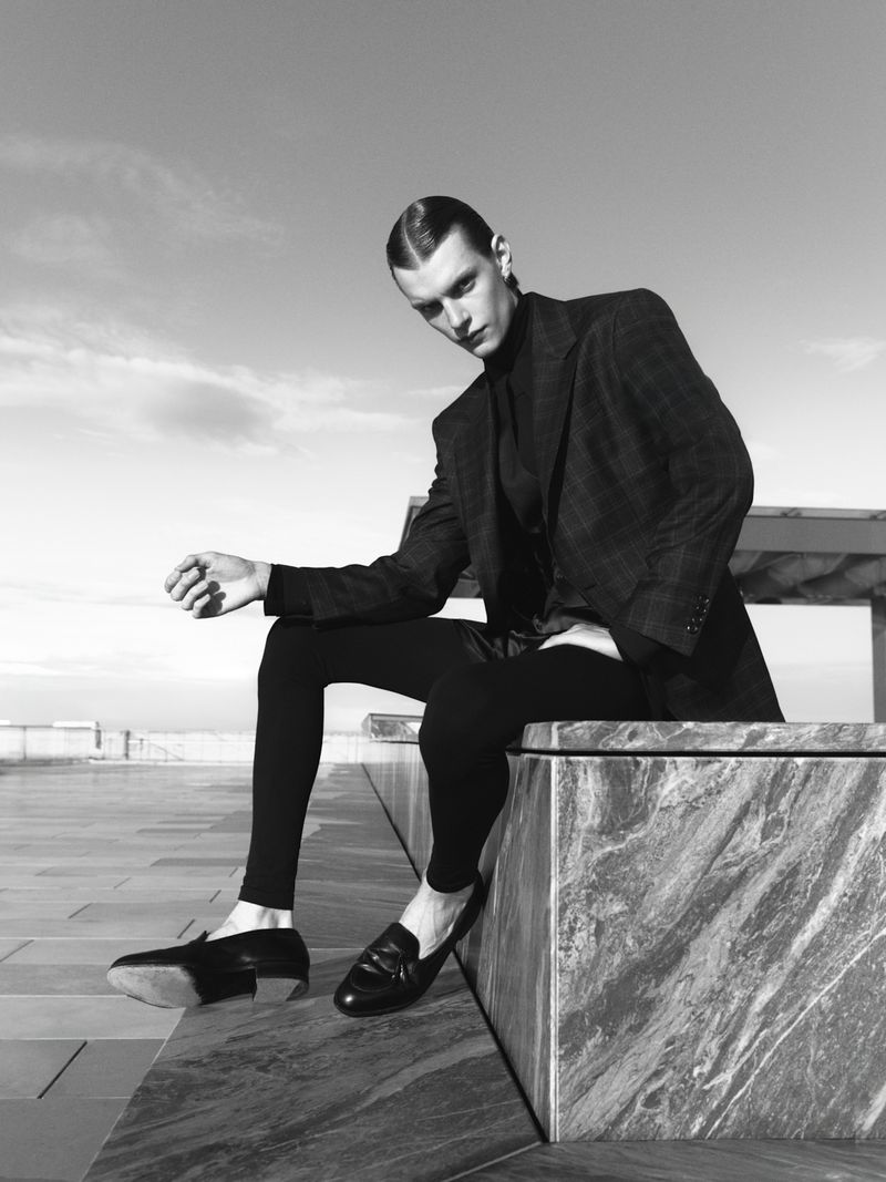 Monochrome portrait of a stylish adult male model in blazer and leggings seated outside against a sky background, fashion advertising.