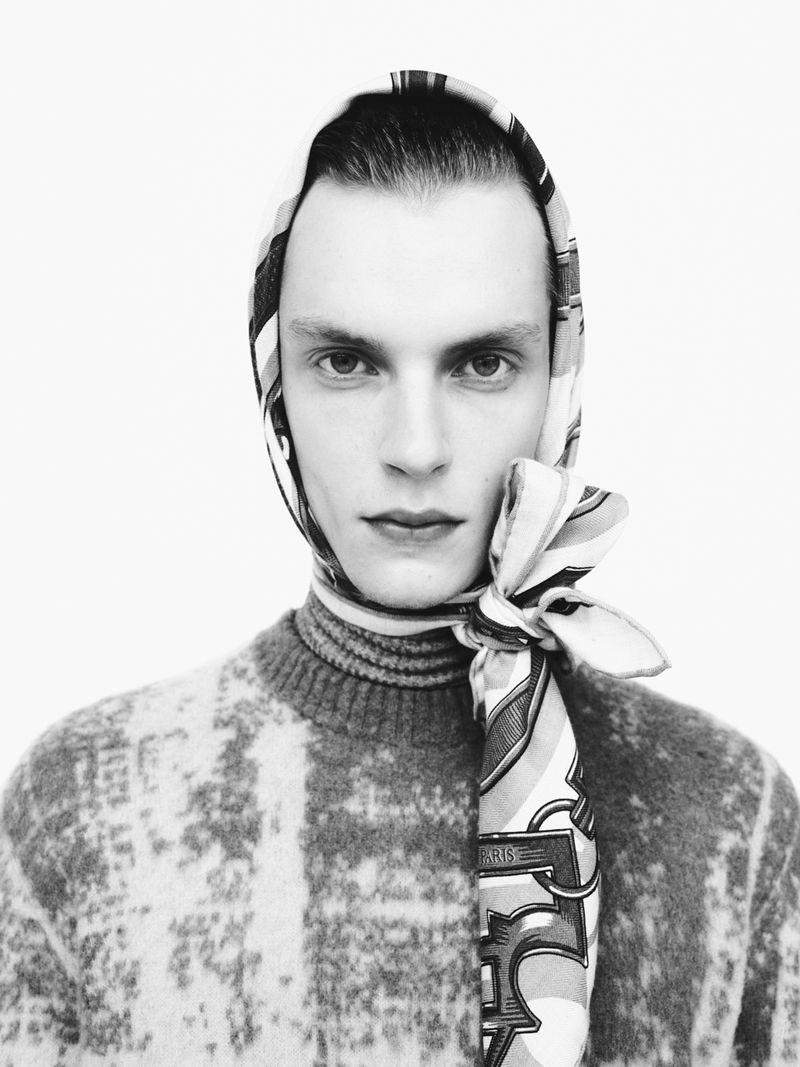 Black and white portrait of a male fashion model wearing a patterned sweater and patterned scarf wrapped around his head indoors.