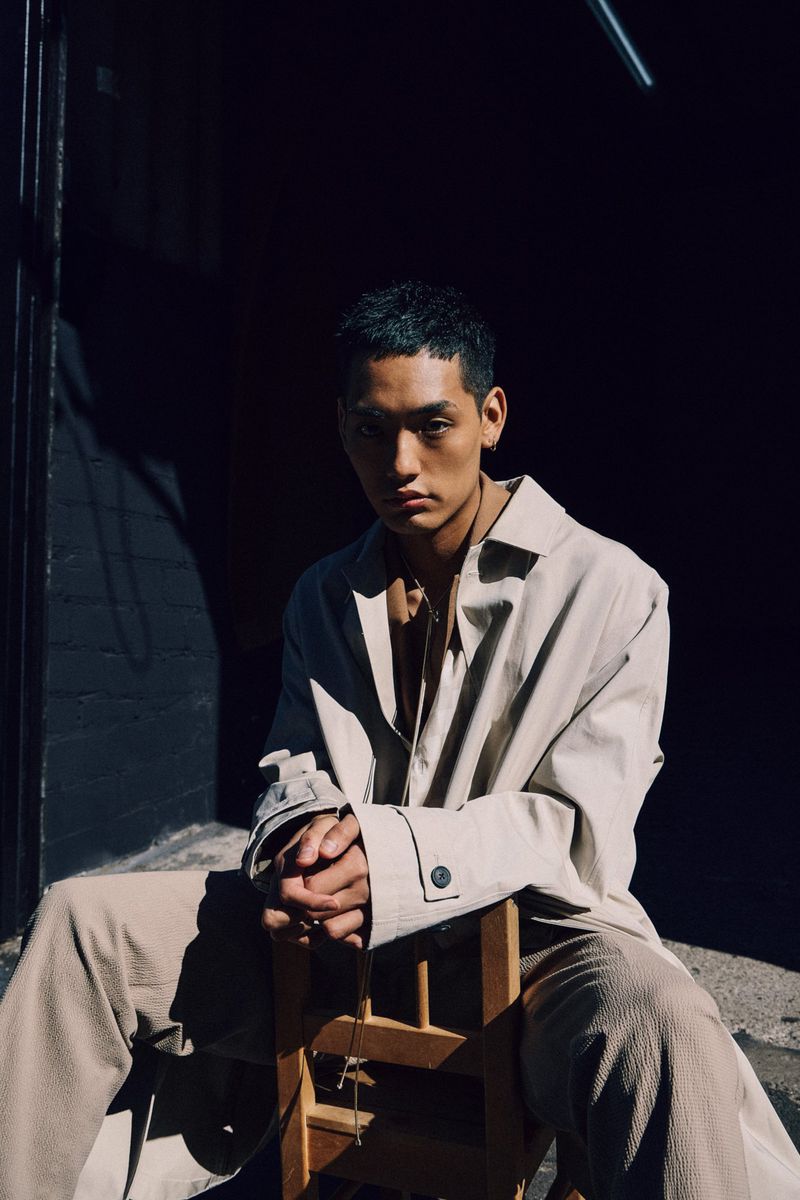Portrait of a young Asian man with dark hair wearing a beige coat and pants, seated on a wooden chair indoors with dark lighting.