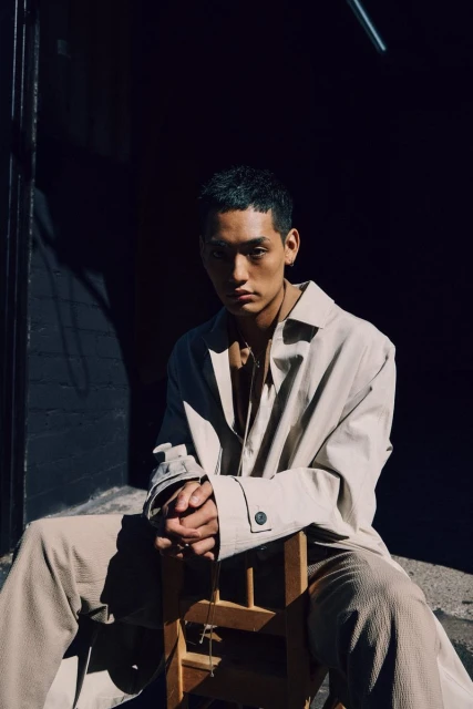 Portrait of a young Asian man with dark hair wearing a beige coat and pants, seated on a wooden chair indoors with dark lighting.