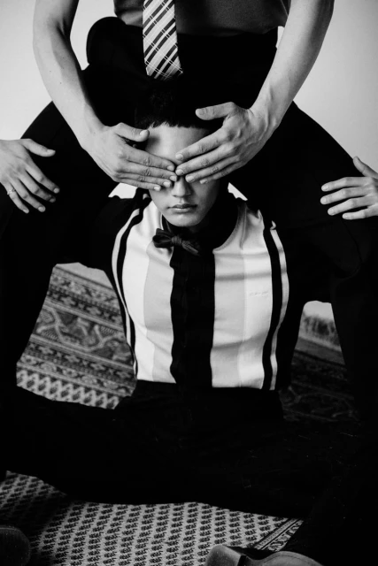 Two young adult men in a black and white portrait, one sitting with the other sitting on their shoulders while covering their eyes.