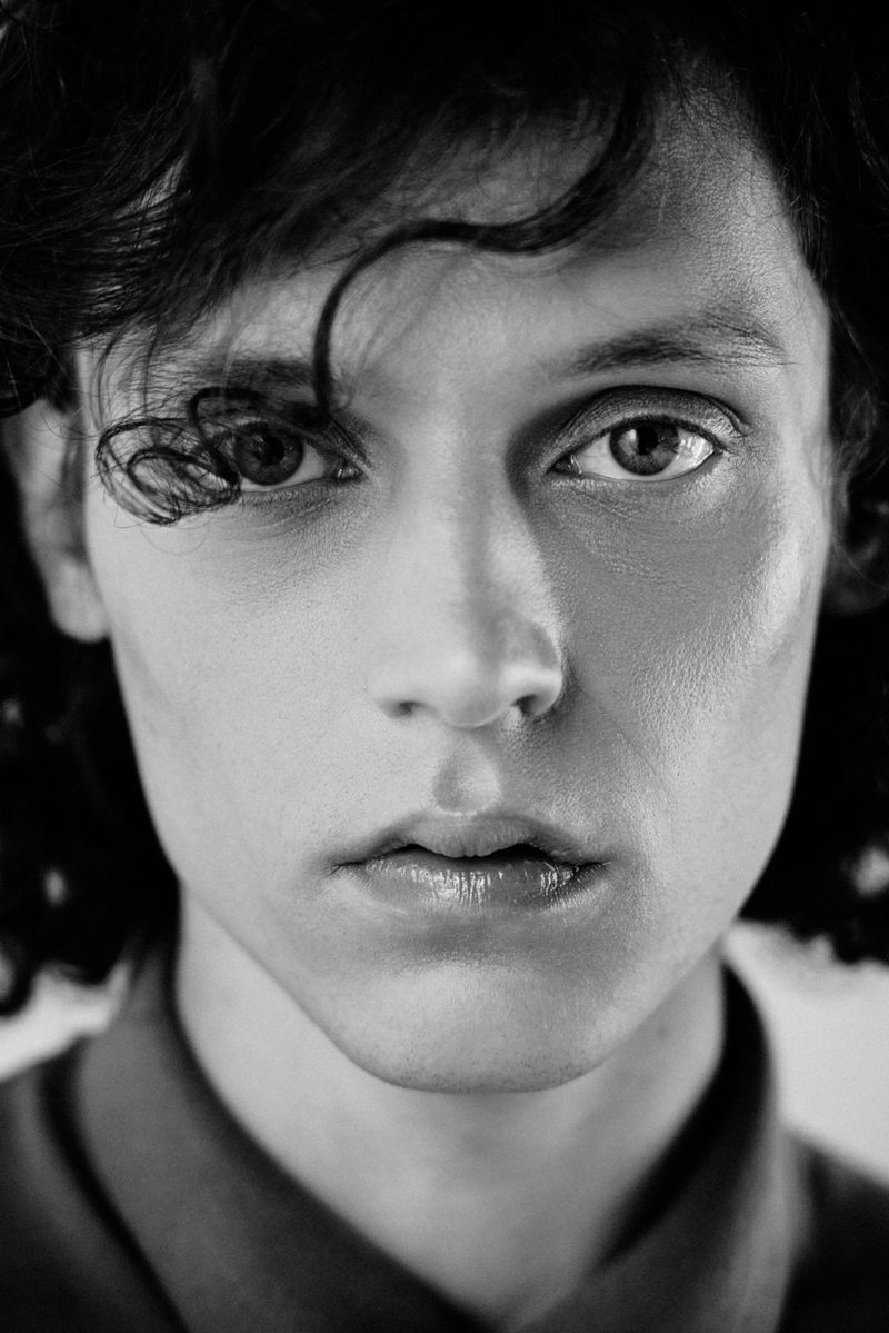 Close-up portrait of a young man with dark curly hair and intense gaze, photographed by Tavinho Costa.