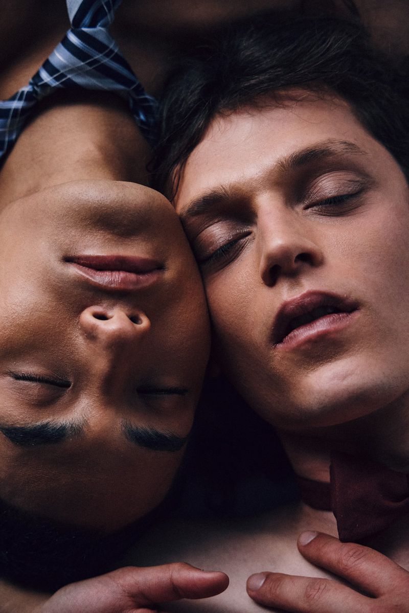 Close-up of two young men with closed eyes, their faces nearly touching, wearing dress shirts and ties in a softly lit, intimate composition.
