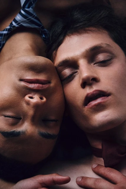 Close-up of two young men with closed eyes, their faces nearly touching, wearing dress shirts and ties in a softly lit, intimate composition.